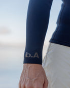 青いサーフィンウェットスーツの袖口に「b.A」のロゴが入った部分のアップ写真。背景は海辺の空